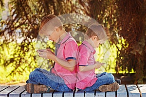 Two little boys playing games on mobile phone in sunny day, sitting back to back.