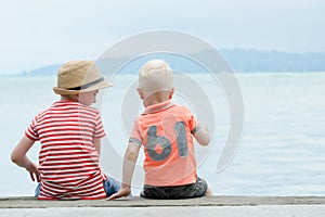 Two little boys sit on a pier, against the sea and mountains. Ba