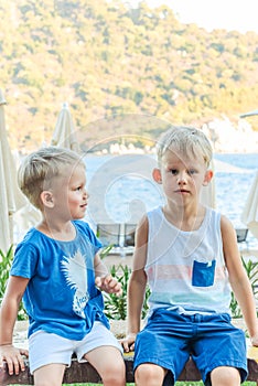 Two little boys are sitting on a bench near a beach laughing and engaging playfully