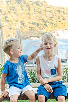 Two little boys are sitting on a bench near a beach laughing and engaging playfully