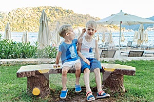 Two little boys are sitting on a bench near a beach laughing and engaging playfully