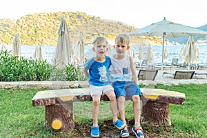 Two little boys are sitting on a bench near a beach laughing and engaging playfully