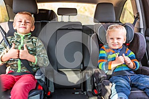 Two little boys sitting on a car seat and a booster seat buckled up in the car