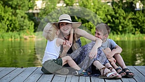 Two little boys whisper something in their mom`s ear. Summer day. Pier on the river bank. Happy family