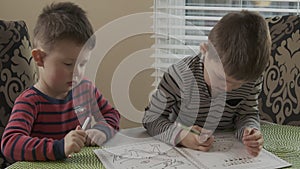 Two little brothers are sitting at the table and draw together on the same coloring. The older brother teaches thhappy