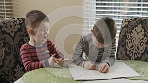 Two little brothers are sitting at the table and draw together on the same coloring. The older brother teaches thhappy