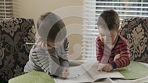 Two little brothers are sitting at the table and draw together on the same coloring. The older brother teaches thhappy