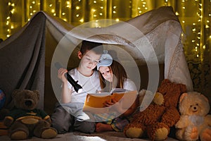 Two little child play at home in the evening to build a camping tent to read books with a flashlight and sleep inside