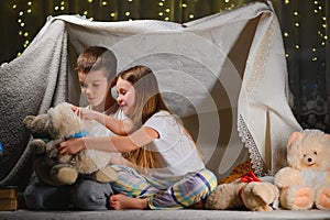 Two little child play at home in the evening to build a camping tent to read books with a flashlight and sleep inside
