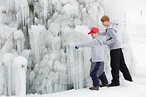 Two little child play under frozen waterfall, exploring the strange ice formations.