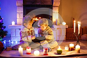 Two little children sitting by a fireplace at home on Christmas time. Happy cute adorable toddler boys, blond twins
