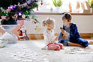 Two little chilren, cute toddler girl and school kid boy playing together card game by decorated Christmas tree. Happy