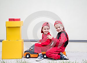Two little courier boys in red uniforms are sitting on a large toy car with yellow cardboard boxes on a light background