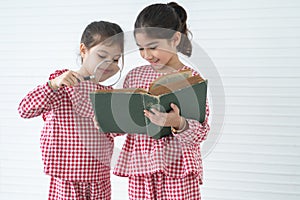 Two little cute children girls, reading a book with magnifying glass while standing on white background. Siblings relationship and