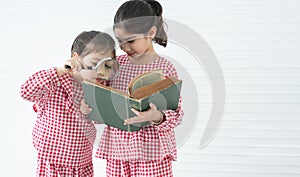 Two little cute children girls, reading a book with magnifying glass while standing on white background. Siblings relationship and