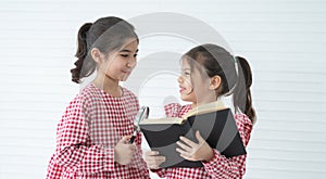 Two little cute children girls, reading a book, playing with magnifying glass while standing on white background. Siblings