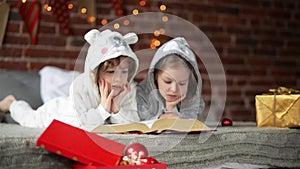 Two little cute siblings reading a book in bed near Christmas tree with lights and illumination. Happy family of two