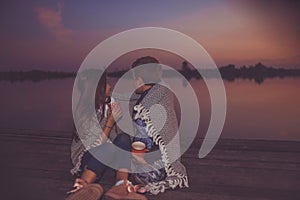 Two little friends, boy and girl talking and drinking tea while sitting covered with blanket by the river. sunset