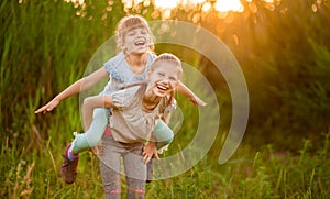 Two little funny sister playing outside on sunset. Little girl climbed up on back of cute sister and fly