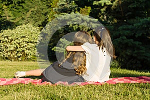 Two little girlfriends embracing sitting on a meadow in the park. View from the back