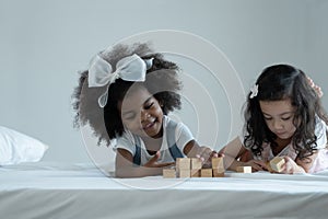 Two little girls, African and Asian kids enjoy playing build wooden block together on bed at bedroom