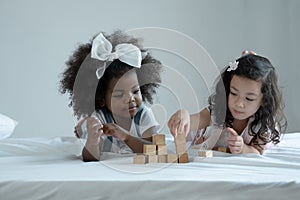 Two little girls, African and Asian kids enjoy playing build wooden block together on bed at bedroom