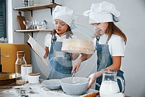 Two little girls in blue chef uniform preparing food by using sieve on the kitchen and reading receipt book