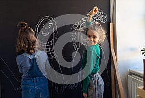 Two little girls drawing with chalks on blackboard wall indoors in playroom, looking at camera.