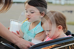 two little girls are drinking milk shake from motherâ€™s hands while sitting in a big black toy car