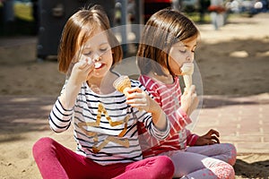 Two little girls eats ice-cream while sitting on the playground