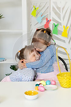 two little girls embrace at a table with a basket of Easter eggs.