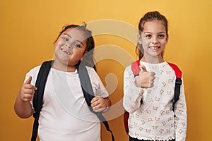 Two little girls friends wearing student backpack smiling happy and positive, thumb up doing excellent and approval sign