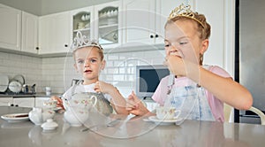 Two little girls having a tea party at home. Sibling sister friends wearing tiaras while playing with tea set and having