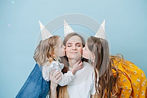 Two little girls kissing their mother on a cheek, all in party cones over blue