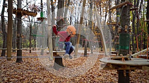 Two little girls in orange helmets and protective gear on rope-way in autumn forest. Children are engaged climbing rope park.