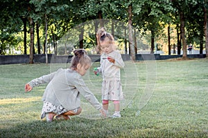 Two little girls playing in park on grass with soap bubbles on warm summer day