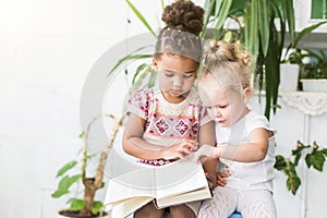 Two little girls read a book on the background of plants in pots