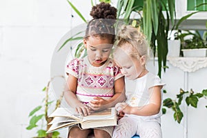 Two little girls read a book on the background of plants in pots