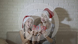 Two little girls in Santa`s hats are holding the red numbers 2019, sitting together on the couch, dancing and smiling