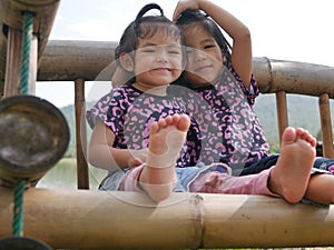 Two little girls, sisters, 2 and 3 years old, smiling and making a heart shape on a bamboo bench near a lake - sisters bond & love