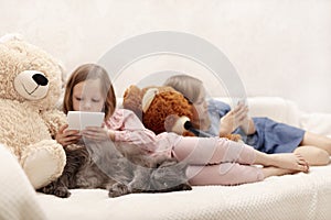 Two little girls sisters 7 and 8 years old are watching an e-book and a smartphone at home on the sofa with their cat