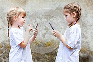 Two little girls, sisters or friends using their smartphones, children texting on their mobile phones facing each other