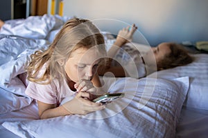 Two little girls sisters are playing with their smartphones lying on a bed in a large hotel room. the harm of gadgets