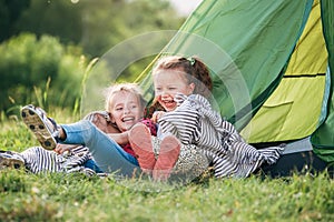 Two little girls sisters sitting on the green grass next to camping tent entrance, cheerfully laughing about jokes they told to