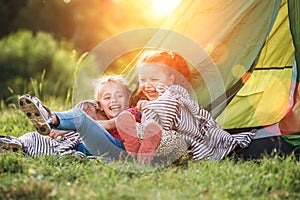 Two little girls sisters sitting on the green grass next to camping tent entrance, cheerfully laughing about jokes they told to