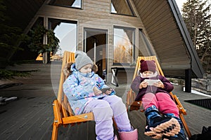 Two little girls sit on chairs at terrace off grid tiny house in the mountains and watching cartoons on mobile phones