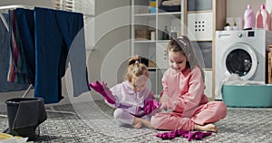 Two little girls sit on the laundry room floor. The sisters are putting pink rubber gloves on their hands, getting ready