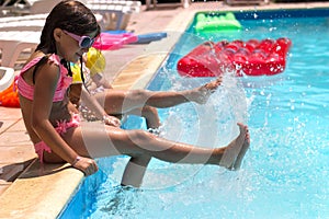 Two little girls sitting on the poolside, playing, splashing water; summertime background