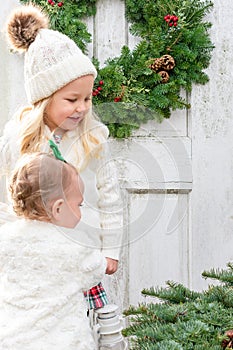 Two little girls in white jackets standing in front of a rustic white door in winter