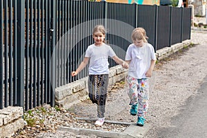 Two little happy school age girls, cheerful children jumping, running down the street alone holding hands and smiling, road side